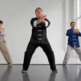 Tre personer utför qi gong i en ljus lokal. De står med böjda knän och armarna i olika positioner.