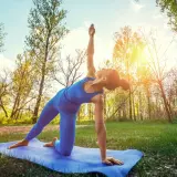 En kvinna gör yoga utomhus i en skog. Solen skiner genom träden.