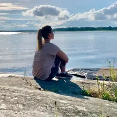 Kvinna sitter på en klippa vid havet, njuter av lugnet. Perfekt för mental avslappning och att ladda batterierna för att sova gott.