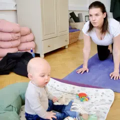 En kvinna i vit topp och svarta leggings gör baby yoga på en lila matta. En bebis sitter på en filt med leksaker.