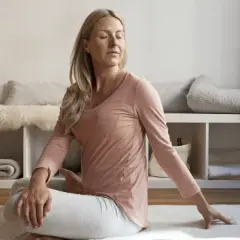 Kvinna i avslappning mindfulness-position på en matta. Bakom henne syns en hylla med filtar och kuddar.