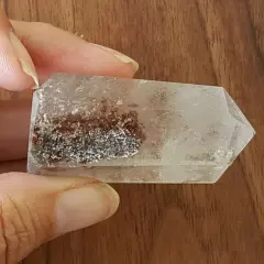 En hand håller en klar kristall med inneslutningar av rödbruna och silverfärgade mineraler, som visar lodolitens fascinerande egenskaper.