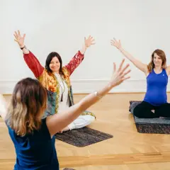 Tre kvinnor deltar i en medicinsk yoga utbildning. De sitter på mattor med armarna lyfta.