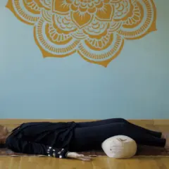 En kvinna vilar på en matta med ögonbindel, en del av en yoga nidra utbildning. En Buddha-staty och korglampor skapar en lugn atmosfär.