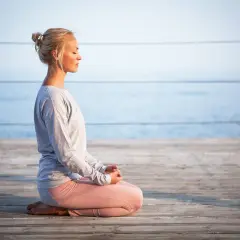 Kvinna med slutna ögon sitter i meditationsställning på en träbrygga vid havet. Hon utstrålar mental avslappning.