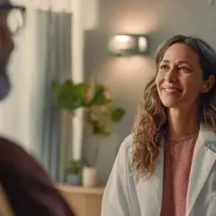 Läkare ler mot patient. Hon har en vit rock och en rosa tröja. Bakom henne syns en datorskärm med grafer, en växt och en infusionsställning. Detta kan vara en del av en funktionsmedicin utbildning.