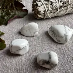 Fyra vita stenar med grå ådring, troligen magnesit, ligger på en grå duk. En bunt salvia och mörka blad ligger bredvid. Magnesit egenskaper kan vara lugnande.