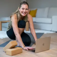 Glad kvinna förbereder sig för ett hatha flow yoga-pass hemma, med en laptop och yogablock redo.