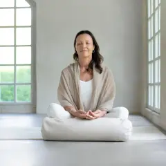 Kvinna i meditationsställning med slutna ögon, omgiven av ljus från stora fönster. Perfekt för guidad meditation text.