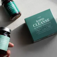 En hand håller en burk med "Rapid Cleanse Morning" kosttillskott, bredvid en låda och en burk för natten. Produkter för en intensiv rening, inspirerade av ayurvedisk medicin.