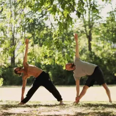 Två män utövar yoga i en park. De står i en triangelställning under trädens grönska.