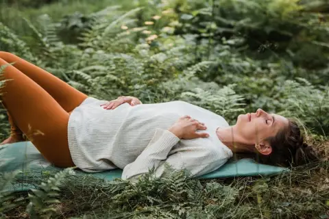 Kvinna ligger på en matta i skogen, med slutna ögon och händerna över bröstet, i en djup avslappning under yoga nidra.