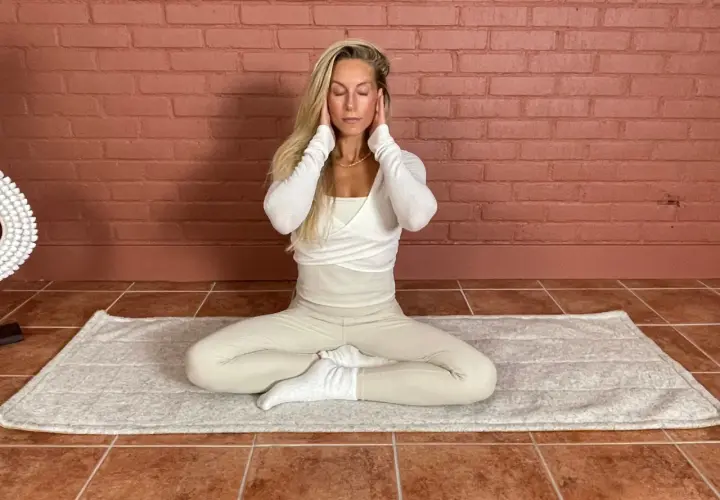 Kvinna med slutna ögon sitter i meditationsställning på en matta. Hon har händerna vid tinningarna. Detta kan vara en del av en lymfyoga utbildning.