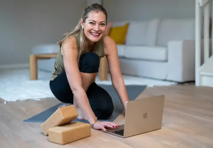 Glad kvinna förbereder sig för kundaliniyoga positioner hemma, med en laptop och yogablock redo.