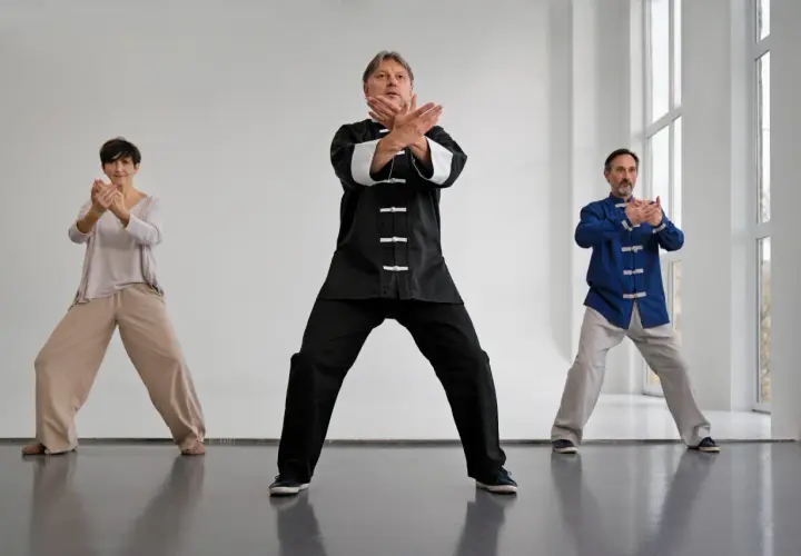 Tre personer utför qi gong i en ljus lokal. De står med böjda knän och armarna i olika positioner.