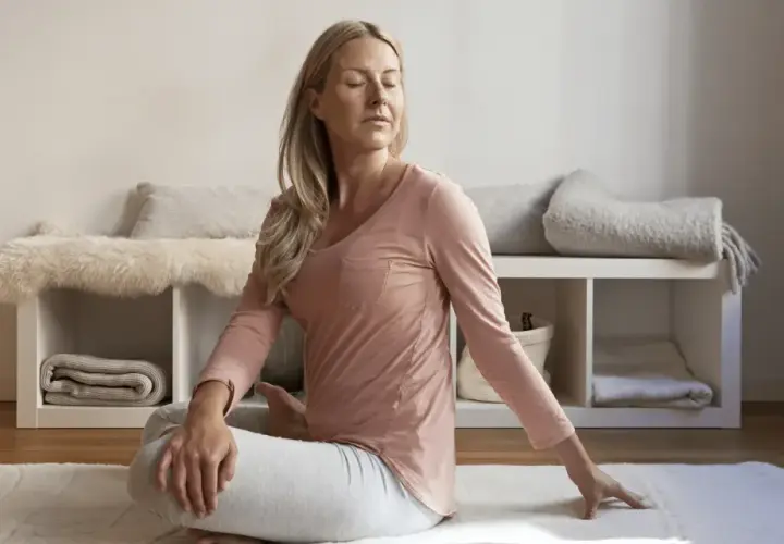 Kvinna i avslappning mindfulness-position på en matta. Bakom henne syns en hylla med filtar och kuddar.