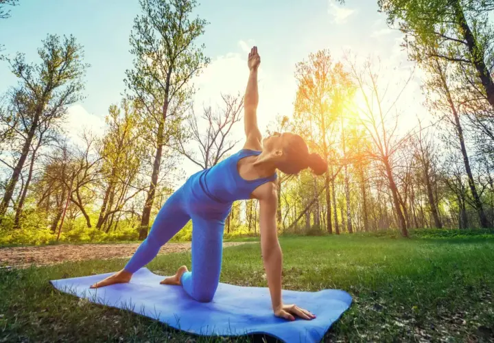 En kvinna gör yoga utomhus i en skog. Solen skiner genom träden.