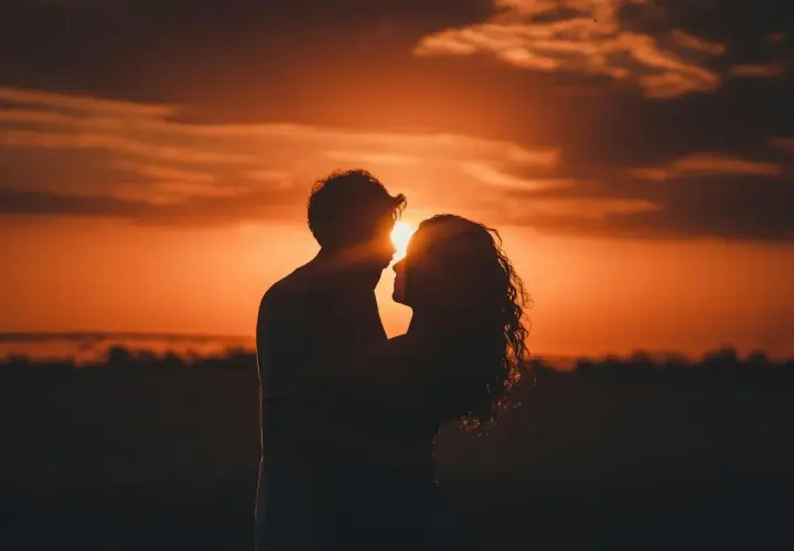 Ett par omfamnar varandra vid solnedgången, solen lyser mellan dem. En våg av kärlek sköljer över dem, lika vacker som himlens våg av färger.