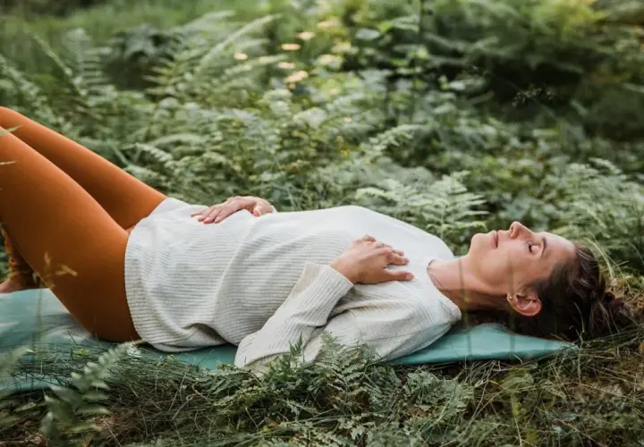 Kvinna ligger på en matta i skogen, med slutna ögon och händerna över bröstet, i en djup avslappning under yoga nidra.