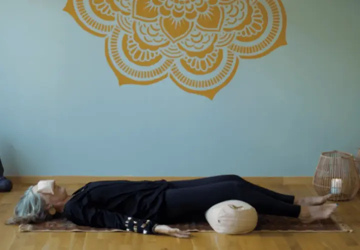 En kvinna vilar på en matta med ögonbindel, en del av en yoga nidra utbildning. En Buddha-staty och korglampor skapar en lugn atmosfär.