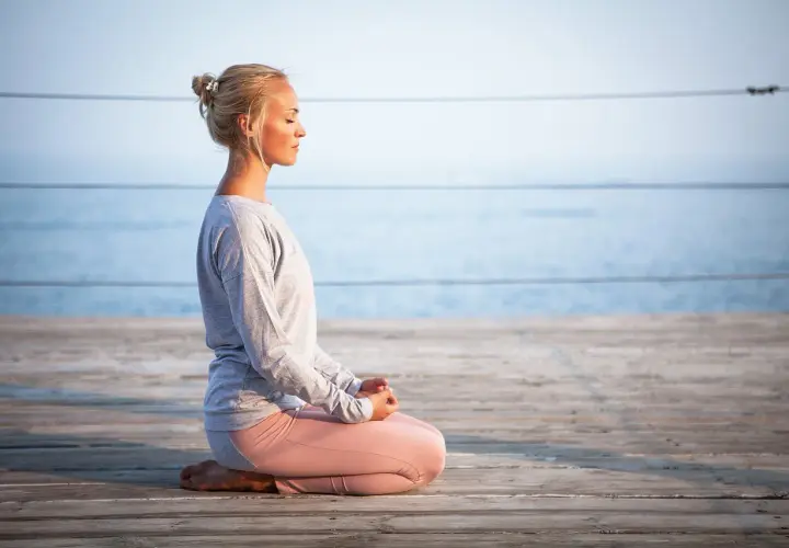 Kvinna med slutna ögon sitter i meditationsställning på en träbrygga vid havet. Hon utstrålar mental avslappning.