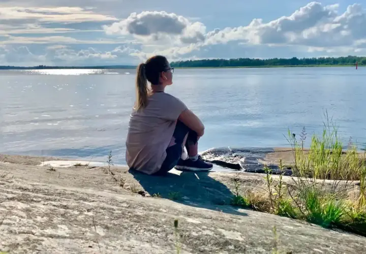 Kvinna sitter på en klippa vid havet, övar mindfulness för att minska stress. Solen glittrar på vattnet.
