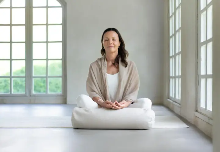 Kvinna i meditationsställning med slutna ögon, omgiven av ljus från stora fönster. Perfekt för guidad meditation text.