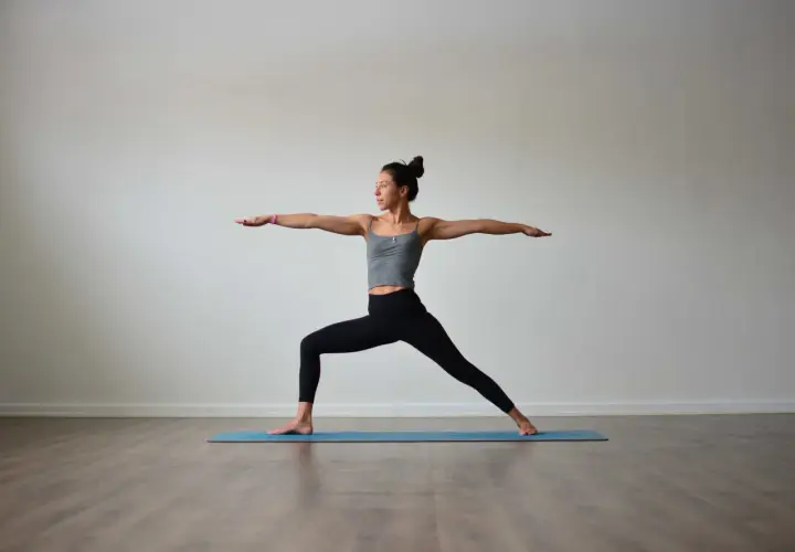 Kvinna i krigare II-ställning under vinyasa yoga. Hon står på en blå yogamatta med armarna utsträckta.