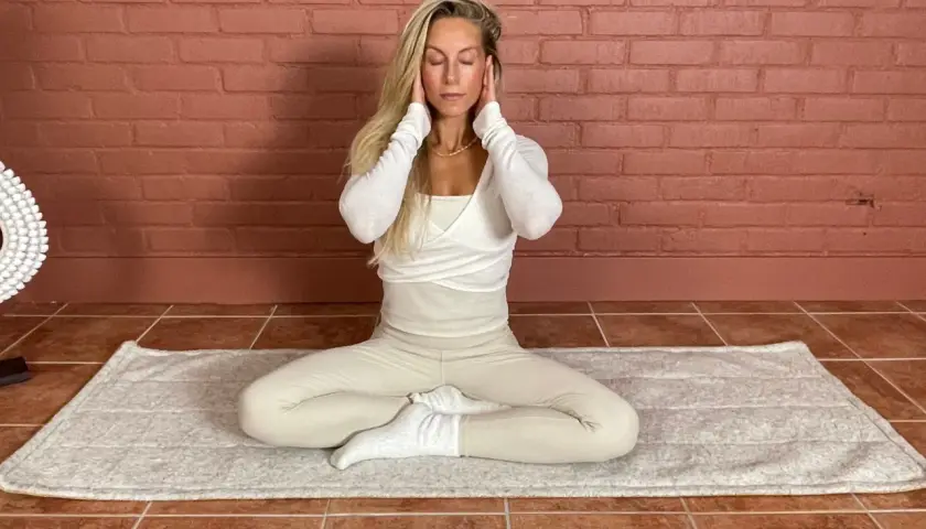 Kvinna med slutna ögon sitter i meditationsställning på en matta. Hon har händerna vid tinningarna. Detta kan vara en del av en lymfyoga utbildning.