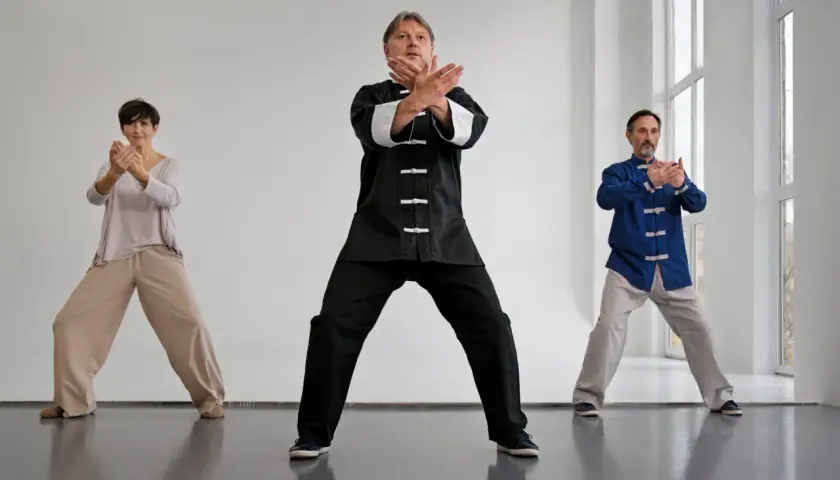 Tre personer utför qi gong i en ljus lokal. De står med böjda knän och armarna i olika positioner.