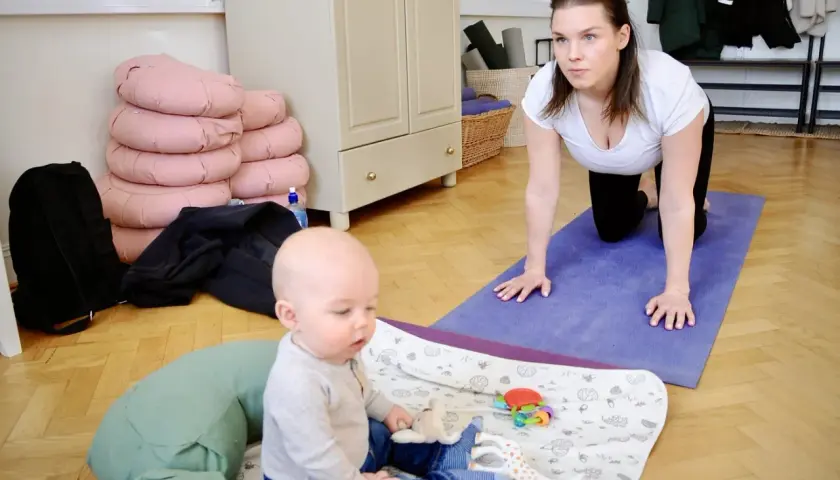 En kvinna i vit topp och svarta leggings gör baby yoga på en lila matta. En bebis sitter på en filt med leksaker.