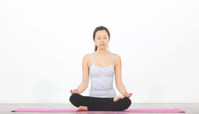 En kvinna sitter i lotusställning på en rosa yogamatta, ögonen slutna, och utövar pranayama yoga.
