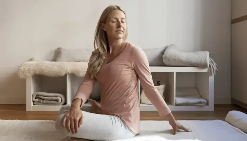 Kvinna i avslappning mindfulness-position på en matta. Bakom henne syns en hylla med filtar och kuddar.