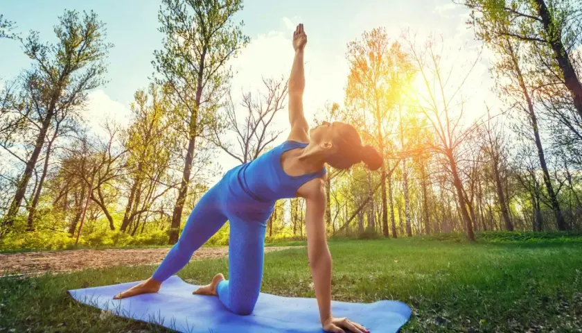 En kvinna gör yoga utomhus i en skog. Solen skiner genom träden.