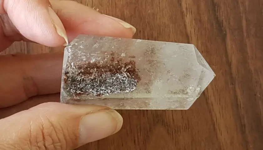 En hand håller en klar kristall med inneslutningar av rödbruna och silverfärgade mineraler, som visar lodolitens fascinerande egenskaper.