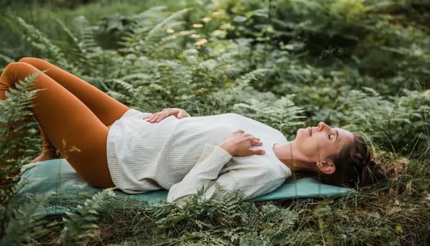 Kvinna ligger på en matta i skogen, med slutna ögon och händerna över bröstet, i en djup avslappning under yoga nidra.