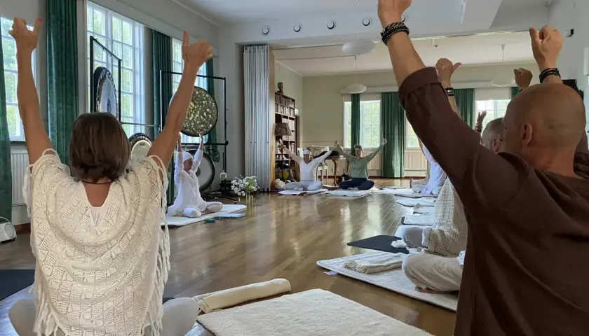 Människor sitter på mattor och höjer armarna under en kundalini yoga-klass. Gongs hänger på väggen.