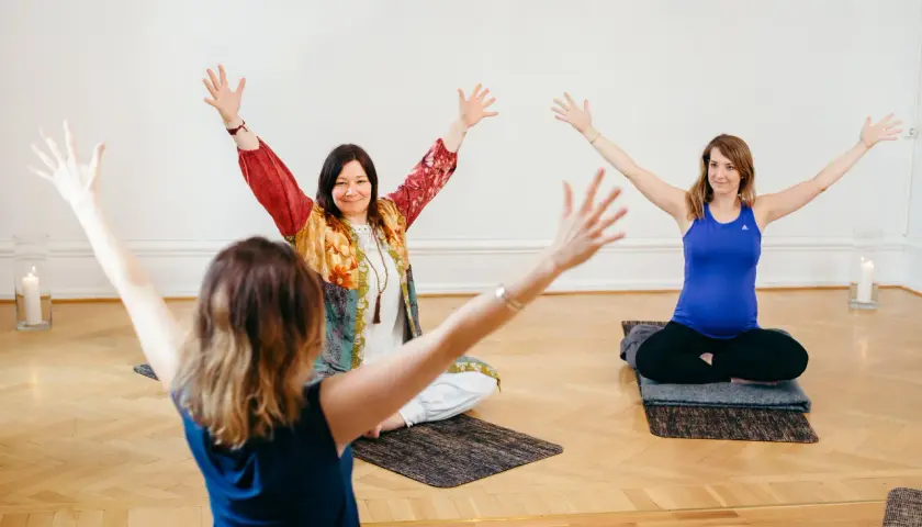 Tre kvinnor deltar i en medicinsk yoga utbildning. De sitter på mattor med armarna lyfta.