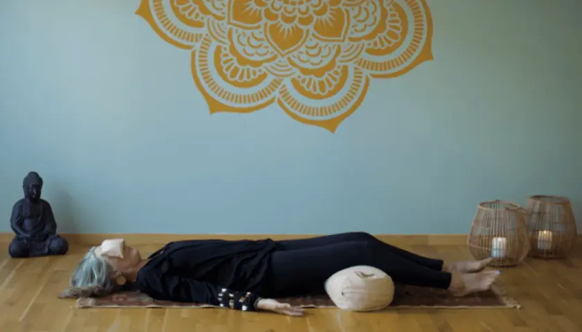 En kvinna vilar på en matta med ögonbindel, en del av en yoga nidra utbildning. En Buddha-staty och korglampor skapar en lugn atmosfär.