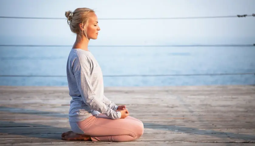 Kvinna med slutna ögon sitter i meditationsställning på en träbrygga vid havet. Hon utstrålar mental avslappning.