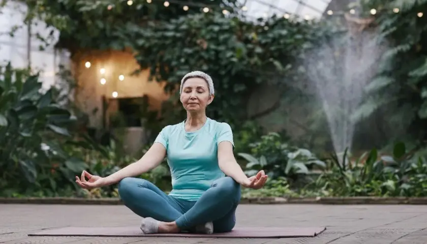 Kvinna i lotusställning, med slutna ögon och händerna i meditation, omgiven av grönska och ljusslingor. En lugn stund för meditation mot stress.