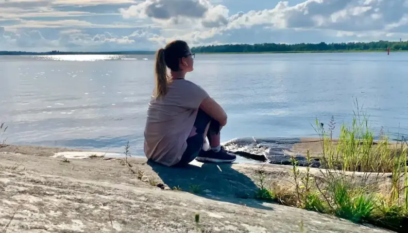 Kvinna sitter på en klippa vid havet, övar mindfulness för att minska stress. Solen glittrar på vattnet.