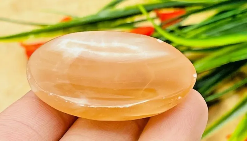 En hand håller en slät, oval honungskalcit. Dess varma, gyllene färg och mjuka lyster framhäver dess unika honungskalcit egenskaper.