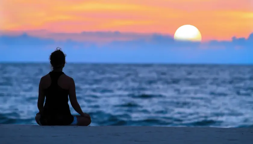 En person sitter i lotusställning på en strand vid solnedgången. En stund av mindfulness i vardagen.