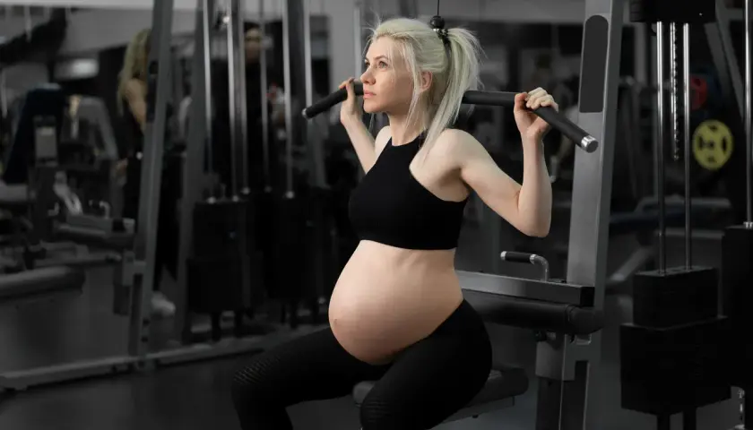 Gravid kvinna i andra trimestern tränar på gym. Hon sitter på en träningsmaskin och drar i ett handtag.