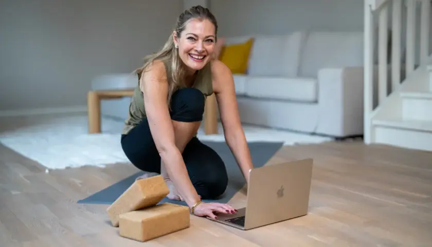 Glad kvinna förbereder sig för ett hatha flow yoga-pass hemma, med en laptop och yogablock redo.