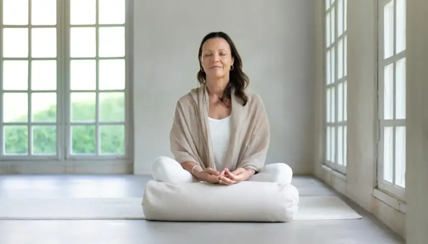 Kvinna i meditationsställning med slutna ögon, omgiven av ljus från stora fönster. Perfekt för guidad meditation text.