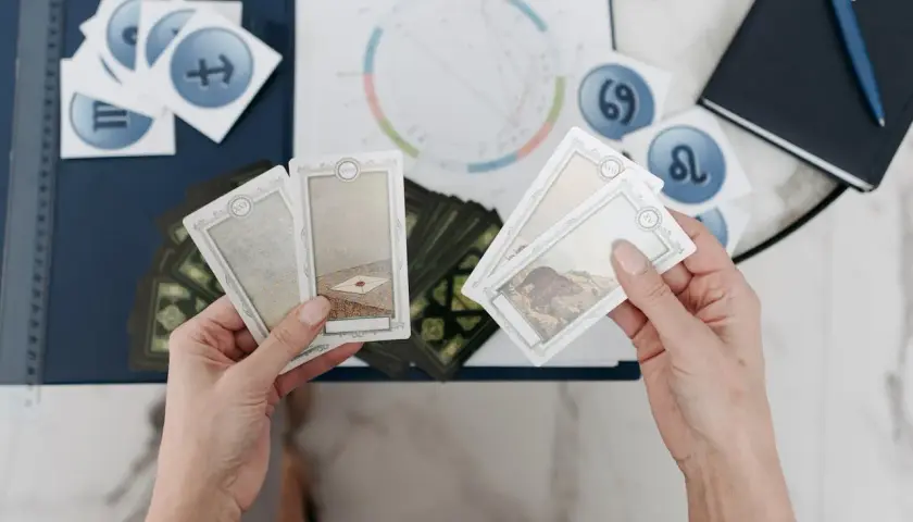 Händer håller tarotkort med symboler för sjunde huset astrologi. En astrologisk karta och stjärntecken syns i bakgrunden.