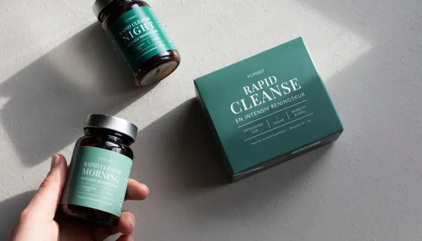 En hand håller en burk med "Rapid Cleanse Morning" kosttillskott, bredvid en låda och en burk för natten. Produkter för en intensiv rening, inspirerade av ayurvedisk medicin.