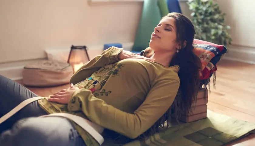 Kvinna vilar med slutna ögon, upplever lugnande yoga nidra effekter. Stöd för huvud och kropp, omgiven av yogatillbehör.